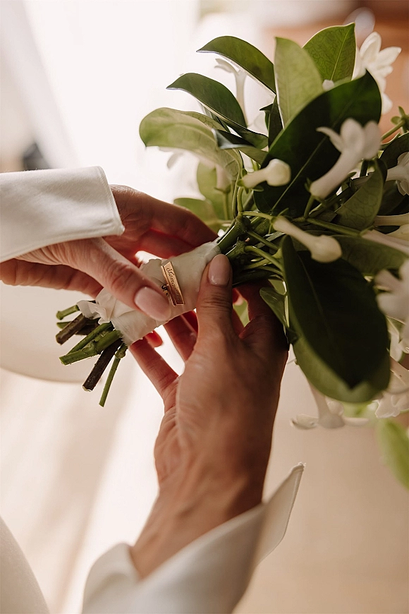Boutonniere pinning on a groom boutonniere, hands fastening white flowers and green leaves with ribbon on a suit jacket in soft indoor light