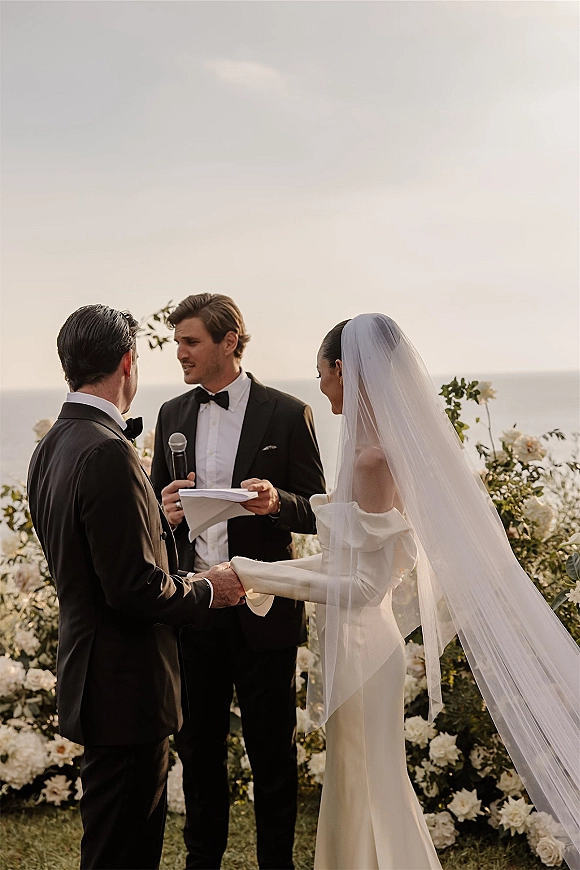 Wedding vows at an outdoor wedding ceremony as bride and groom hold hands, her veil blowing beside an ocean horizon backdrop