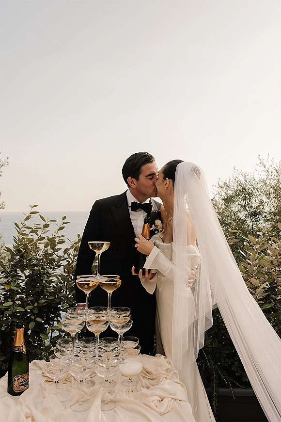 Wedding kiss as bride holds a champagne bottle beside a coupe glass tower, ocean view behind, veil and tuxedo in black-tie style