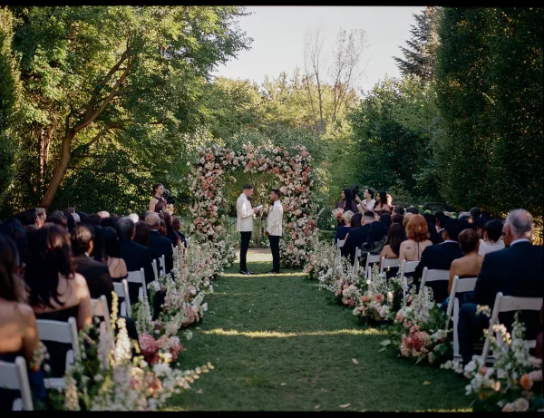 Wedding ceremony with two grooms exchanging vows under a floral arch, guests in white chairs along flower-lined aisle on sunny garden lawn