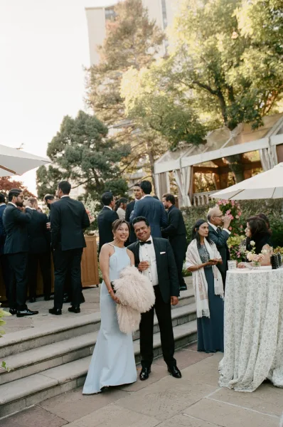 Wedding cocktail hour guests mingle in tuxedos and a light blue dress around high-top tables with florals on a tented terrace