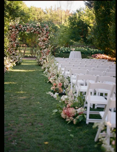 Outdoor ceremony setup with a floral arch and rose-lined aisle, white folding chairs on a sunlit lawn with hedges and trees in back