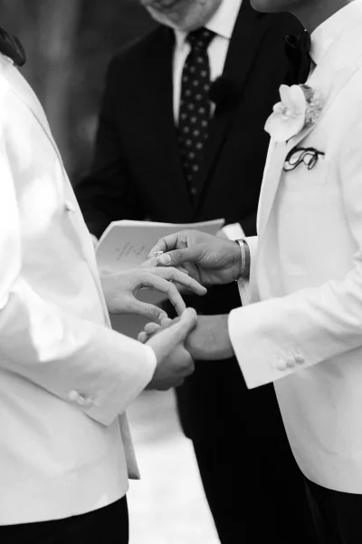 Ring exchange as one groom slides a wedding ring exchange photo moment onto the other’s finger, white tux jackets and officiant blurred behind