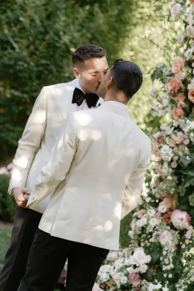 Wedding kiss between two grooms in white dinner jackets under a pastel floral arch with greenery in a garden setting
