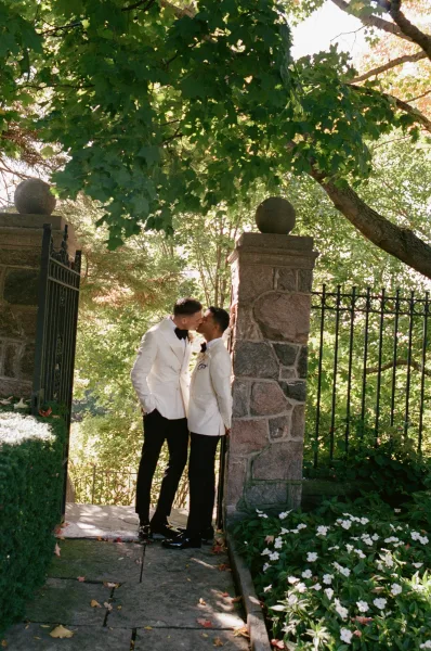 Wedding kiss as two grooms in matching white tuxedos and black bow ties embrace by a sunlit stone gate with garden greenery