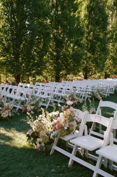 Ceremony aisle decor with white folding chairs, roses and greenery floral clusters lining a sunlit grass lawn beneath trees