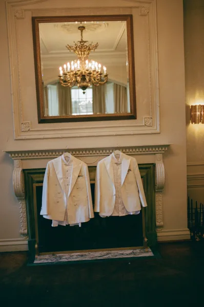Groom suits in white tuxedos with satin lapels hanging beneath a chandelier, displayed against an elegant fireplace and ornate mirror backdrop