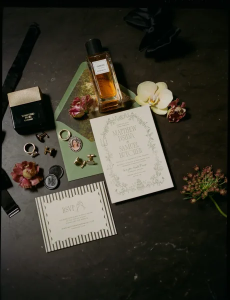 Invitation suite wedding invitation flat lay with RSVP card, sage envelopes, rings, wax seal, pocket watch and orchid sprigs on dark tabletop