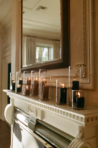 Wedding candle decor with pillar candles in glass hurricane vases arranged along a fireplace mantel beneath a large mirror, paneled wall behind
