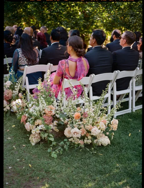 Wedding ceremony guests seated in white folding chairs, with roses and hydrangea aisle florals in a sunlit garden setting
