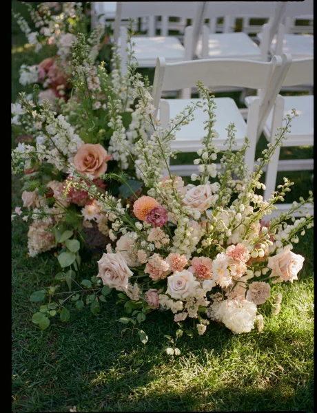 Ceremony aisle flowers in blush and ivory line a grass lawn aisle with roses and hydrangea beside rows of white folding chairs