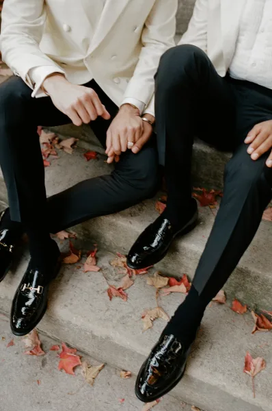 Groom portrait in a white tuxedo jacket and black trousers, seated on stone steps with autumn leaves, showing his wedding ring