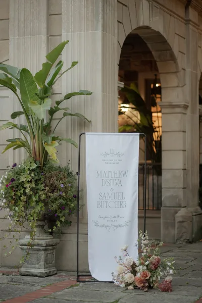 Wedding welcome sign on a black metal frame with a fabric banner and blush florals, set by a stone column and arched doorway entrance