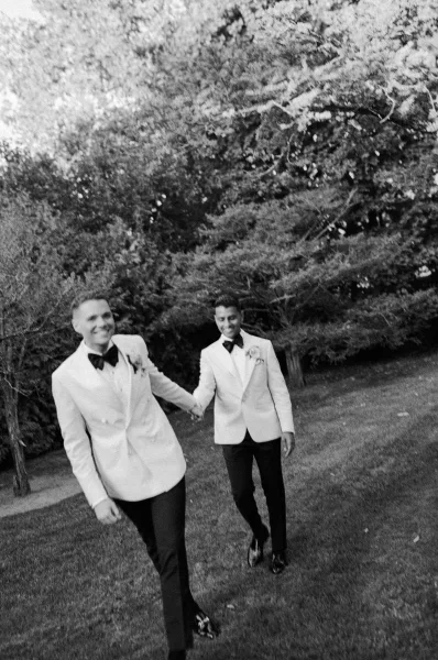 Couple portrait of two grooms holding hands in white tuxedo jackets with boutonnieres, walking on a garden lawn among trees