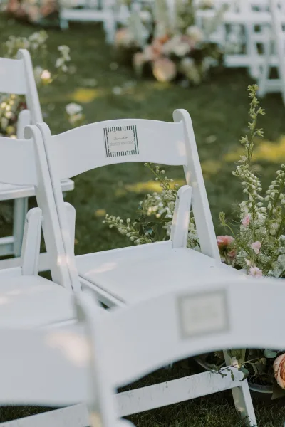 Ceremony seating with outdoor wedding chairs, white folding chairs and a reserved sign beside rose and greenery aisle florals on a lawn