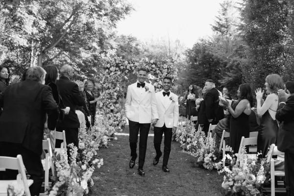 Wedding recessional as two grooms walk down aisle holding hands, in white dinner jackets under a floral arch, guests applauding in garden