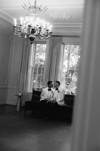 Wedding kiss between two grooms in matching white tuxedos with black bow ties on a sofa beneath a chandelier, tall windows behind