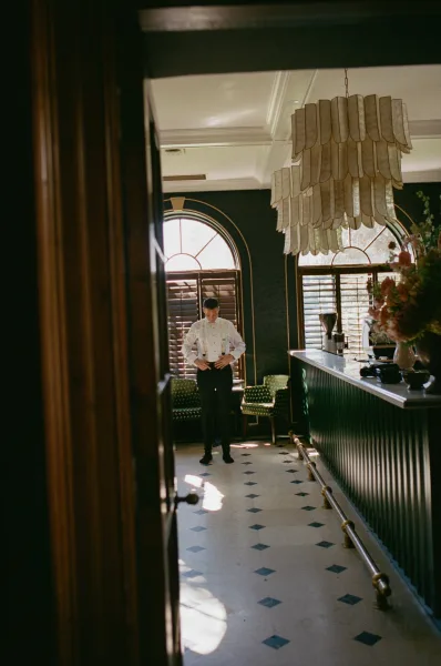 Groom portrait in a white shirt with suspenders, standing in a dark green lounge with arched windows and chandelier, lit by natural light