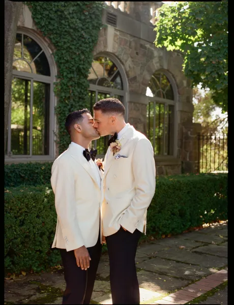 Wedding kiss portrait of two grooms kissing in white tuxedos with black bow ties, framed by an ivy-covered stone building in sunlight