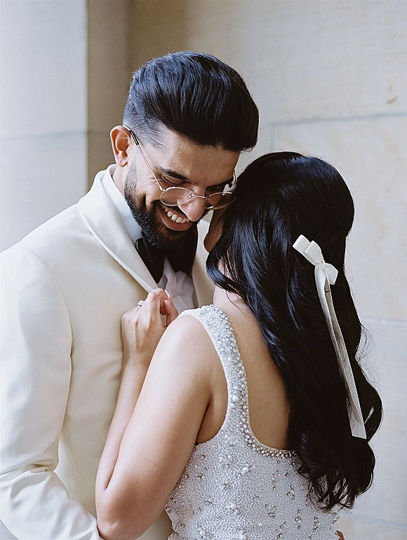 Couple portrait of bride and groom embrace, her beaded wedding dress and hair bow ribbon as he smiles in white tux by a neutral wall