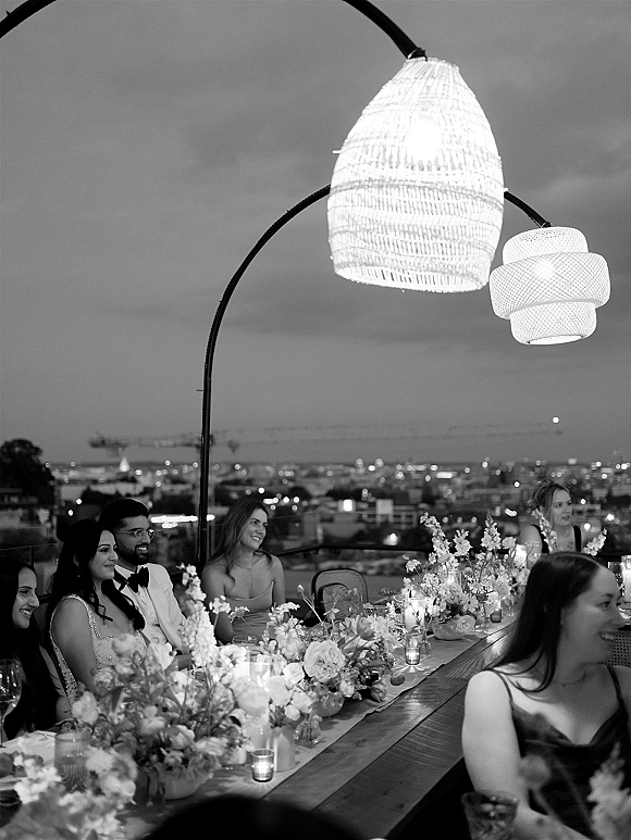 Wedding reception toast on a rooftop wedding reception, with long banquet table lined with taper candles and florals against city skyline at night