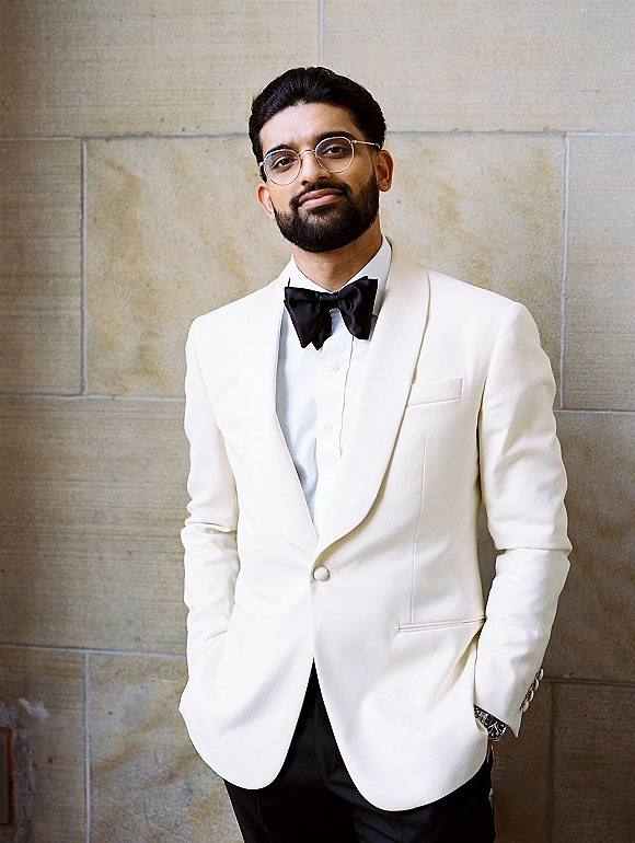 Groom portrait in a white tuxedo groom look with black bow tie and glasses, hands in pockets, standing against a stone wall
