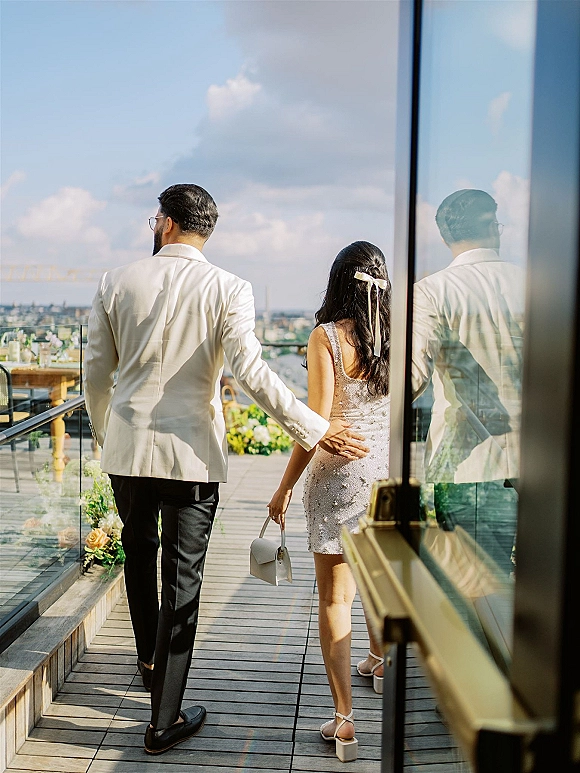 Couple walking together on a sunny rooftop terrace, bride in a short dress with hair ribbon and heels beside groom in white jacket, city skyline beyond glass railing