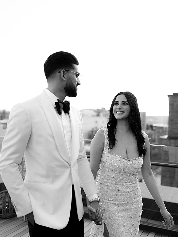 Couple portrait of bride in a beaded wedding dress holding hands with groom in a white tuxedo jacket on a rooftop terrace by the skyline
