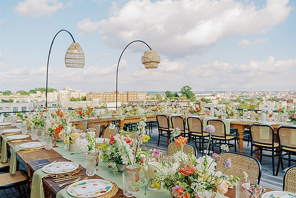 Reception tablescape for a rooftop wedding reception with long wooden tables, pastel florals, taper candles, rattan chargers, skyline backdrop