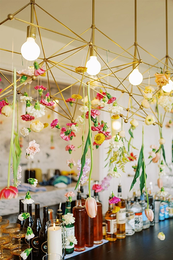 Hanging floral installation with pink and yellow blooms draped from a geometric brass chandelier above an indoor bar with mirrors and glassware