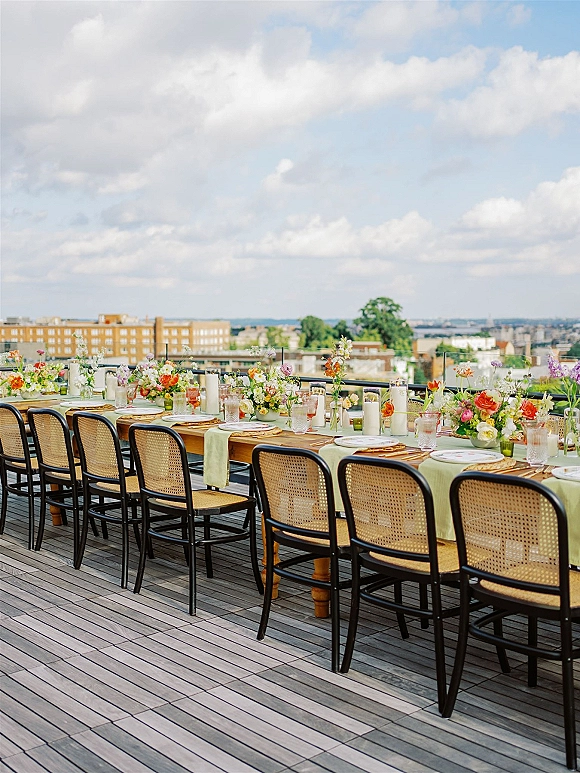 Reception tablescape with a long banquet table, colorful florals and taper candles on a rooftop deck, city skyline beyond the railing