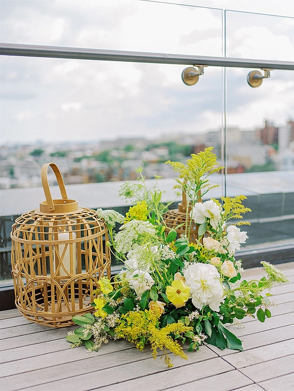 Wedding floral arrangement of yellow and white wedding flowers with a rattan lantern and pillar candle on a rooftop deck overlooking the city skyline