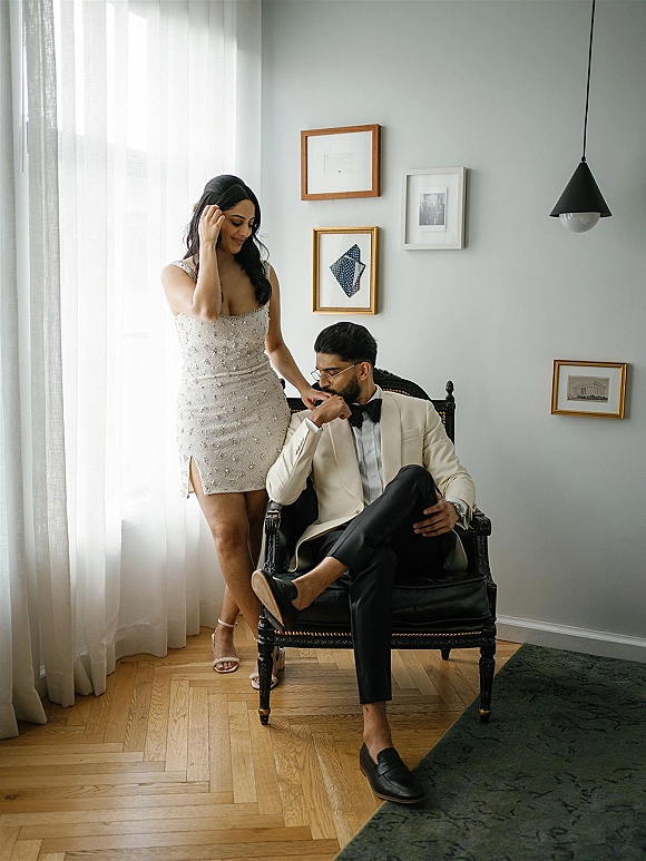 Couple portrait with groom kissing bride hand, bride in a short beaded wedding dress by window light near sheer curtains and armchair