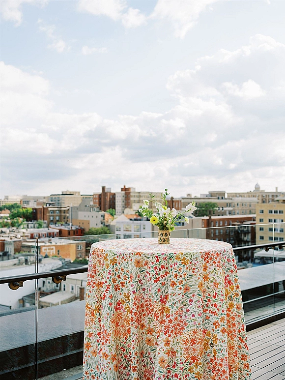 Cocktail table decor with wedding cocktail hour table floral tablecloth and bud vase of yellow-white flowers on a rooftop terrace skyline