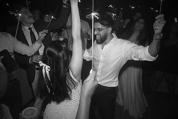 Wedding dance floor packed with guests waving sparklers during a sparkler exit send off in a dark reception room, glasses raised in celebration