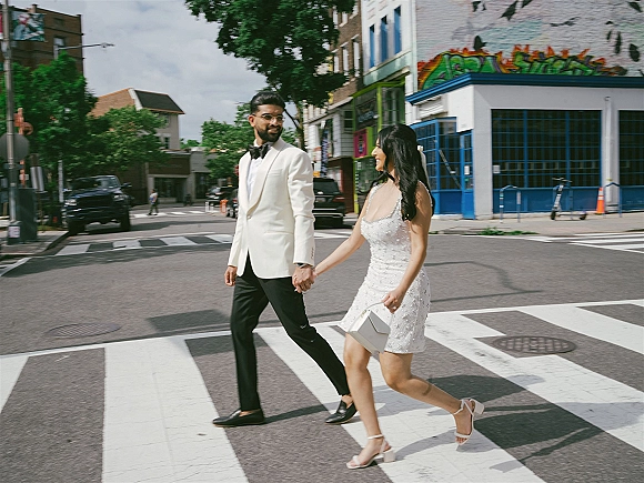 Couple portrait of bride in a short wedding dress and groom in white tuxedo holding hands on a crosswalk by a graffiti mural