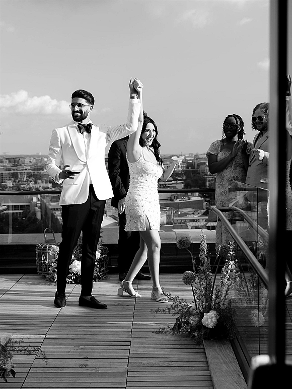 Wedding recessional on a rooftop wedding recessional as newlyweds raise hands, holding champagne coupes on a terrace with skyline backdrop
