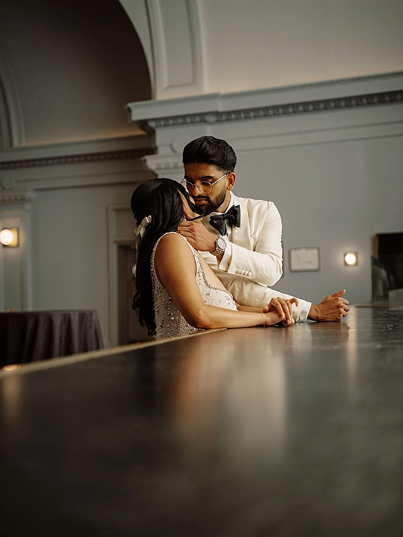 Wedding couple portrait of groom in white tuxedo touching bride’s face as she leans on a bar counter in a moody cocktail lounge setting