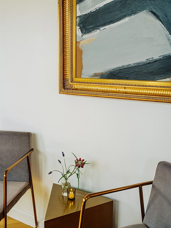 Wedding lounge seating with gray velvet armchairs, gold side table, bud vase with single stem flowers, and framed artwork on white wall