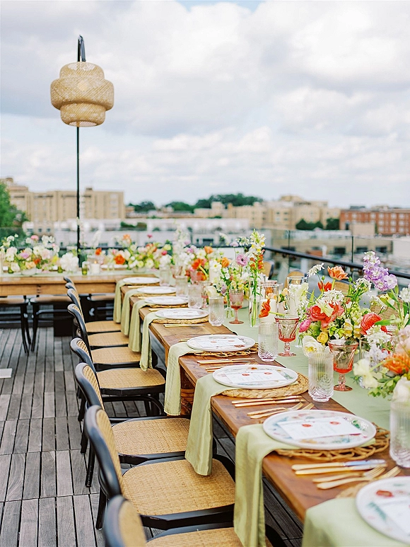 Reception tablescape with a sage green table runner, wildflower bud vases, colored goblets and gold flatware on a rooftop deck overlooking the city skyline