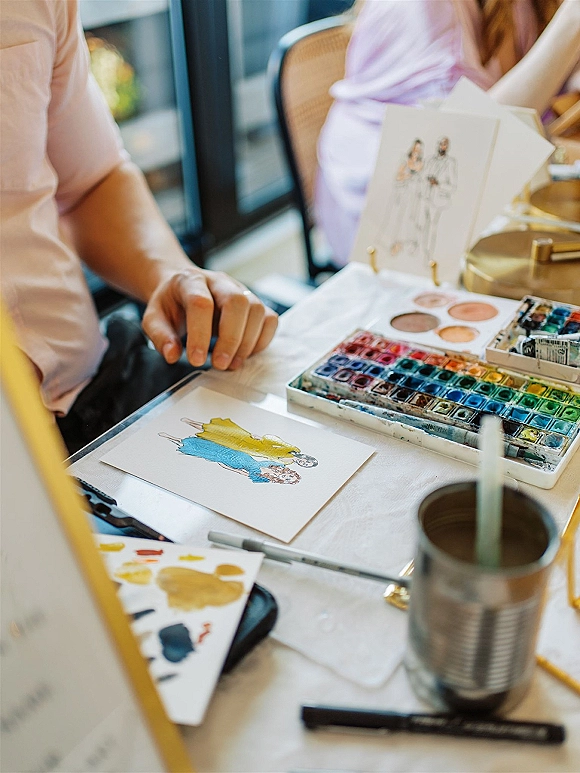 Wedding guestbook illustration station with live wedding painting tools, watercolor palette and brush beside couple card on white tablecloth