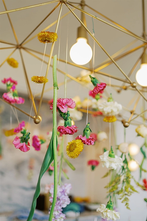 Hanging floral installation with carnations and daisy-like blooms on a gold geometric frame, suspended by string with exposed ceiling light bulbs