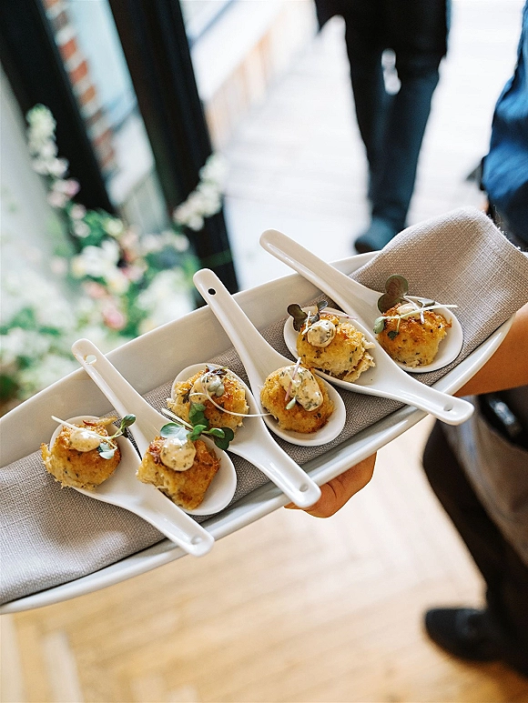 Wedding passed appetizers in white ceramic tasting spoons on a tray with microgreens and sauce, served at an indoor cocktail hour with blurred guests