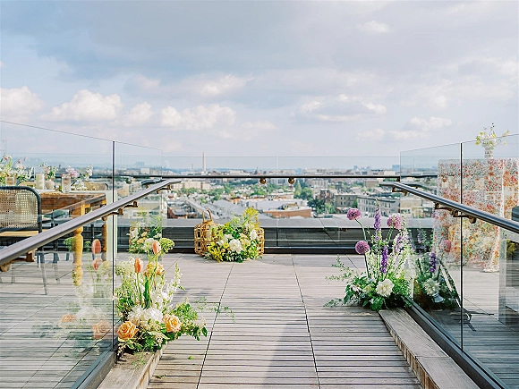 Ceremony aisle decor with ground floral arrangements in wicker baskets beside a glass railing on a rooftop terrace with city skyline backdrop
