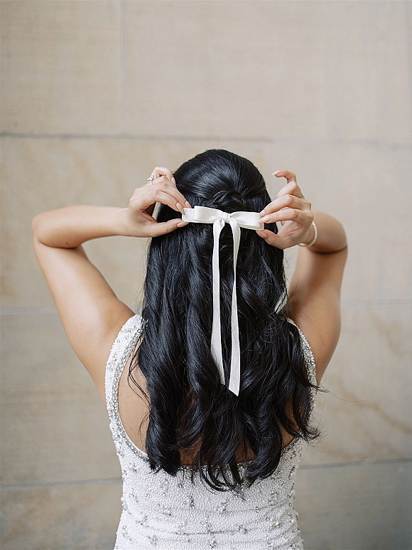 Bridal hairstyle with a wedding hair bow, long wavy half-up hair tied with a white ribbon over a beaded dress back by a stone wall