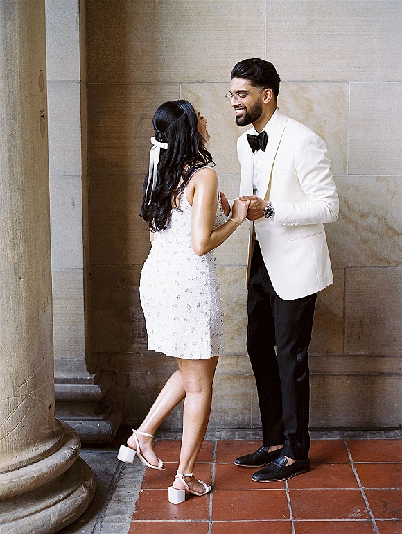 Couple portrait of bride and groom holding hands and laughing, her sequin mini dress and ribbon bow beside stone column and wall