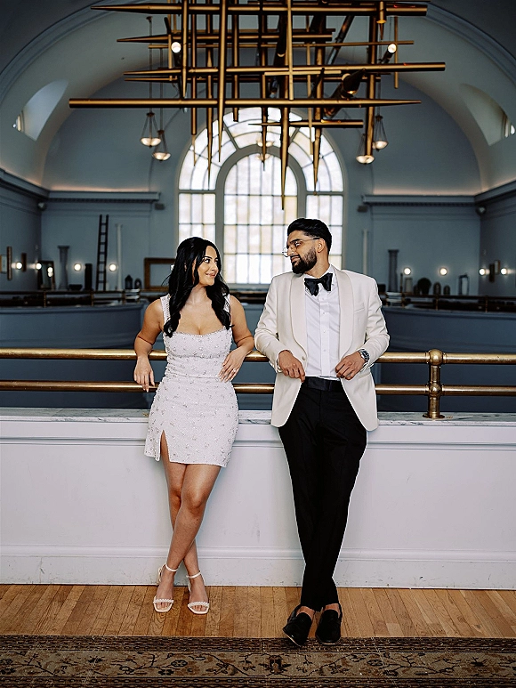 Couple portrait of bride and groom looking at each other, leaning on brass railing under a modern chandelier in a vaulted hall