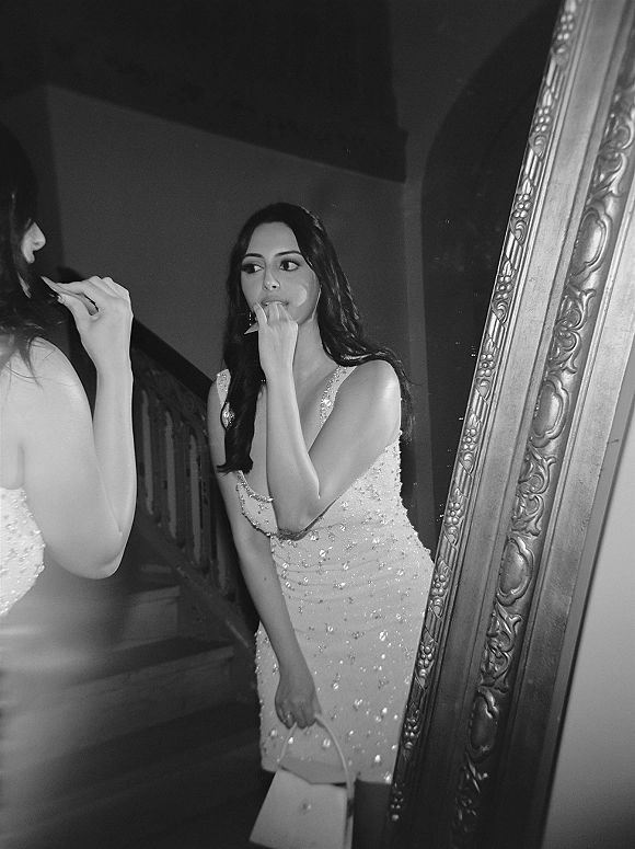Bridal portrait reflected in an ornate mirror, bride in mirror wearing a beaded spaghetti-strap gown, drop earrings, holding a handbag by a staircase