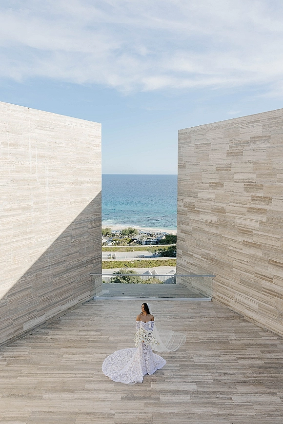 Bridal portrait of a bride in an off the shoulder lace wedding dress holding a white bouquet on a stone terrace with ocean view and blue sky
