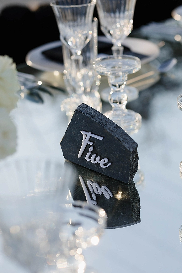 Wedding place card on a slate place card with acrylic name lettering beside crystal glassware and white florals on a reflective table